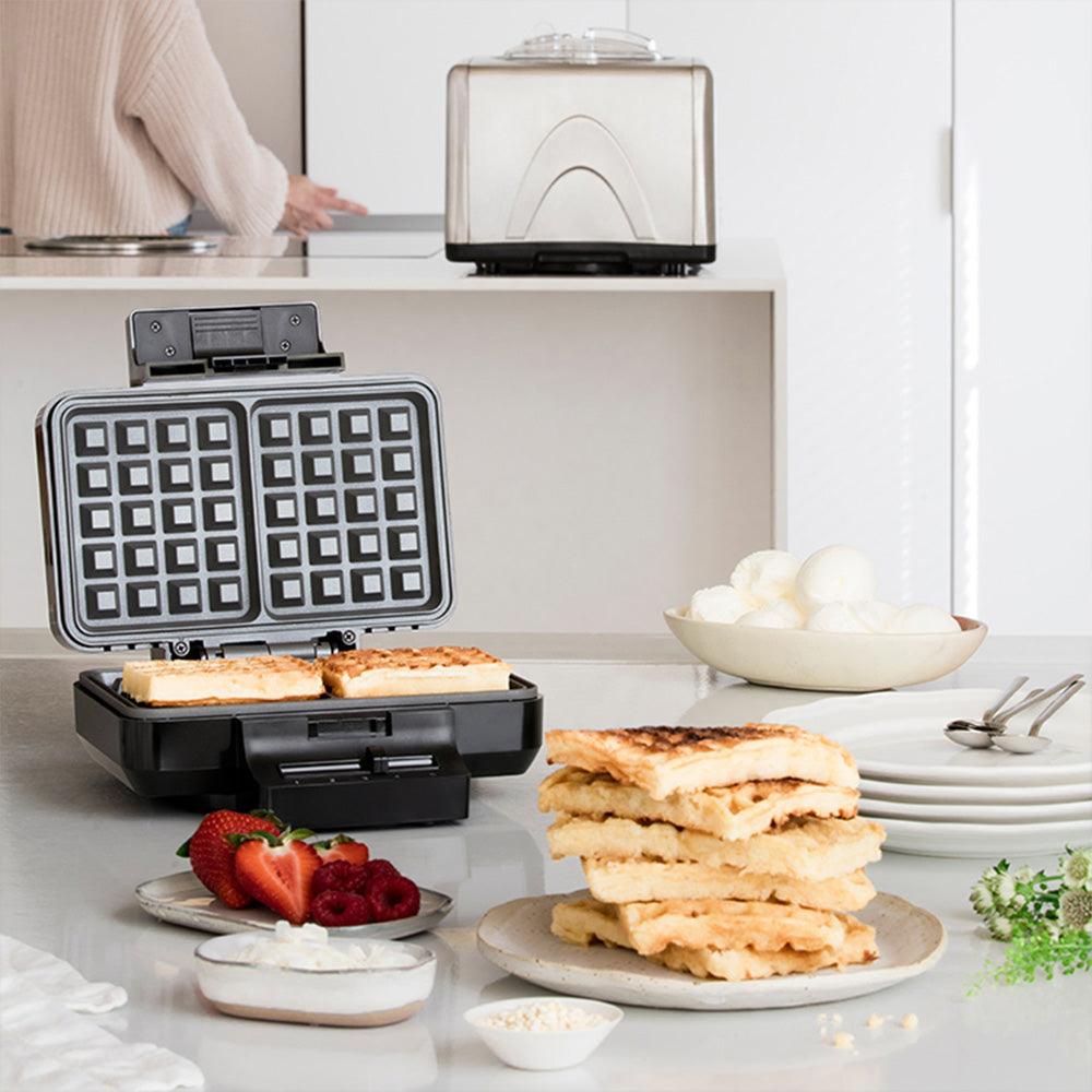 Offenes Waffeleisen auf Küchenarbeitsfläche mit frisch gebackenen Waffeln, serviert mit Beeren, Eis und Toppings für ein einfaches Frühstück zuhause.